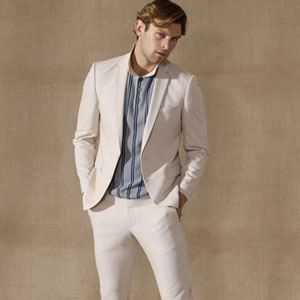 Ein Mann trägt einen eleganten, sandfarbenen Anzug mit einer gestreiften, blauen und weißen Bluse darunter, steht vor einem neutralen Hintergrund.