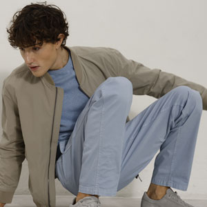 Ein Mann mit lockigem Haar trägt eine beige Jacke und hellblaue Hose, sitzt auf dem Boden und schaut zur Seite.