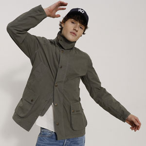 Ein Mann trägt eine graue Jacke mit Taschen, ein weißes T-Shirt, eine blaue Jeans und eine schwarze Kappe, während er eine entspannte Pose einnimmt.