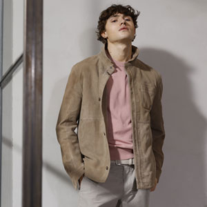 Ein Mann in einer hellbraunen Wildlederjacke und einem rosa Pullover steht in einem Innenraum mit neutraler Wand.