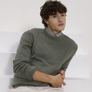 Ein junger Mann mit lockigem Haar trägt einen graugrünen Pullover und sitzt leger auf einem weißen Hintergrund.