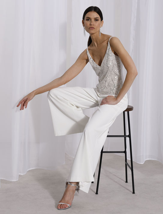 Pflegetipps_ViceVersa_WP Frau in eleganter Kleidung sitzt auf einem Hocker, trägt eine glänzende, silberne ärmellose Bluse und weite, weiße Hosen, in einem minimalistischen Raum mit weißen Vorhängen.