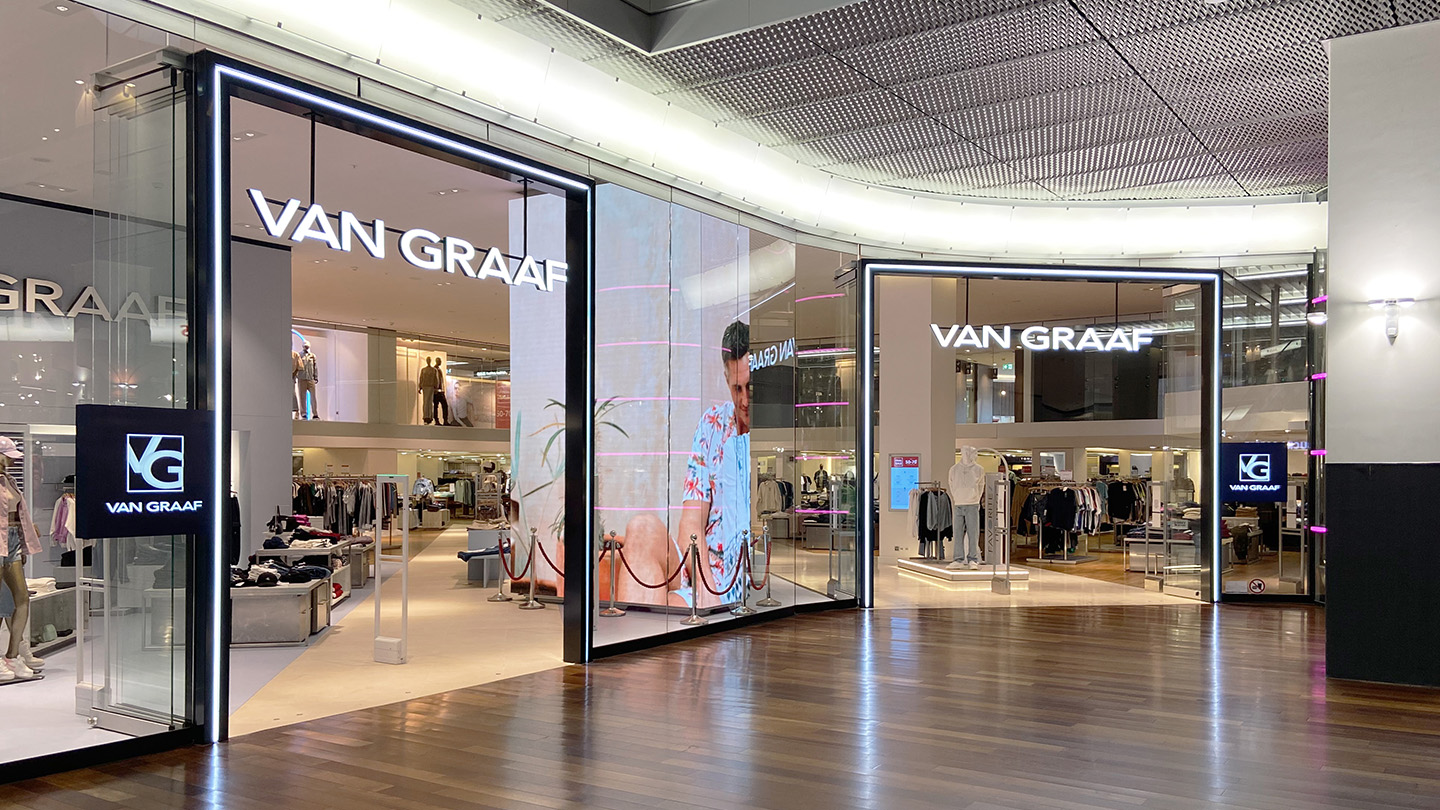 Eingang eines Van Graaf Bekleidungsgeschäfts in einem Einkaufszentrum mit leuchtendem Logo und Sicht auf die Verkaufsfläche im Inneren.