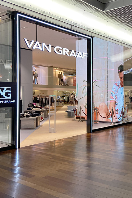 CH_Spreitenbach_Store_TopicSlider Geschäftseingang von Van Graaf mit beleuchtetem Logo und großem Werbebildschirm, der Innenansicht des Ladens im Hintergrund zeigt.