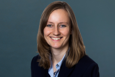 Siri (1) Porträt einer lächelnden Person mit schulterlangem braunem Haar in einem dunklen Blazer und hellen Hemd vor einem neutralen Hintergrund.